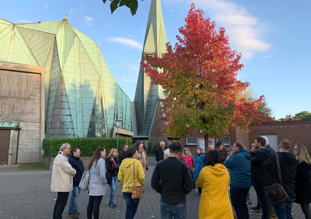 stadt-neuss_ortsmittelpunkte_stadtteilspaziergang-weckhoven-2019-10-10.jpg Eine größere Gruppe Menschen versammelt sich vor einer modernen Kirche und hört einer Person zu. Es ist ein sonniger Tag, im Hintergrund steht ein Baum dessen Blätter sich bereits zur Hälfte herbstlich rot gefärbt haben.